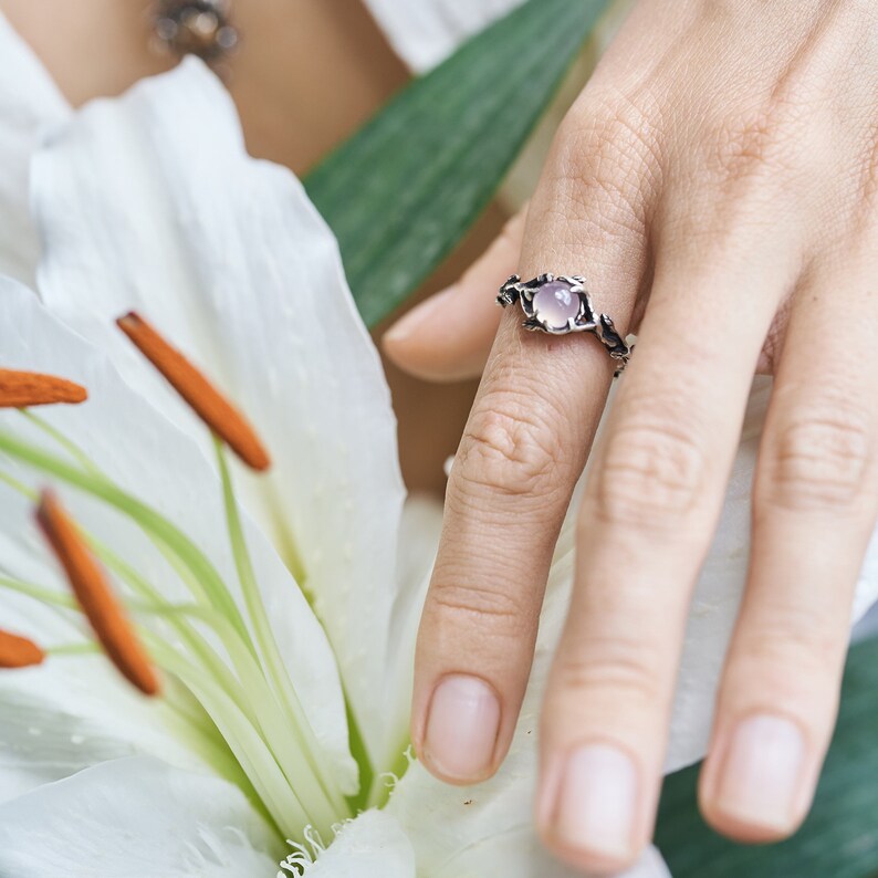May include: A silver ring with a delicate floral design and a pale pink gemstone. The ring is worn on a finger against a backdrop of white flowers.