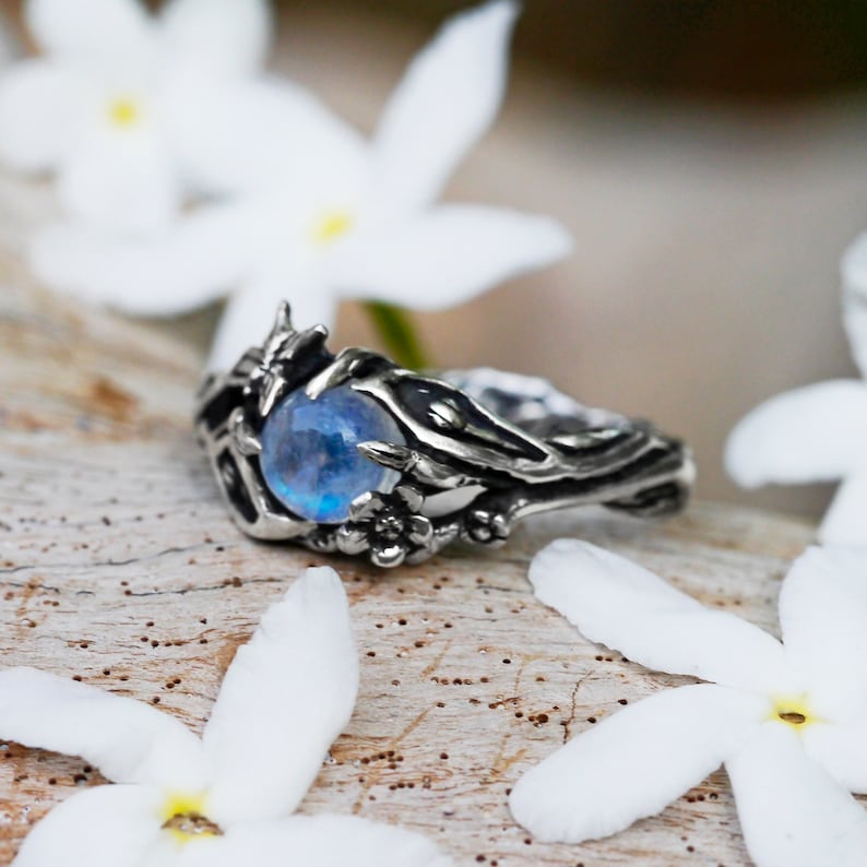 May include: A silver ring with a blue moonstone set in a detailed, organic design resembling branches and leaves. The ring is resting on a wooden surface with white flowers.