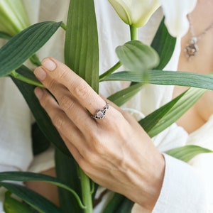 May include: A silver ring with a pink gemstone set in a delicate, twisted band. The ring is worn on a hand holding a white lily.