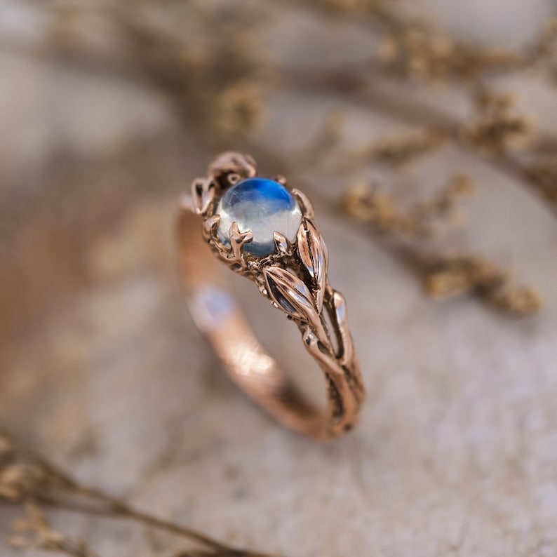 Puede incluir: Un anillo de oro rosa con un delicado dise&ntilde;o de hojas. El anillo presenta una piedra de luna redonda e iridiscente.