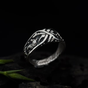 May include: A silver ring with a detailed design featuring a stylized animal figure and intricate lines. The ring has a textured finish and a slightly curved band.
