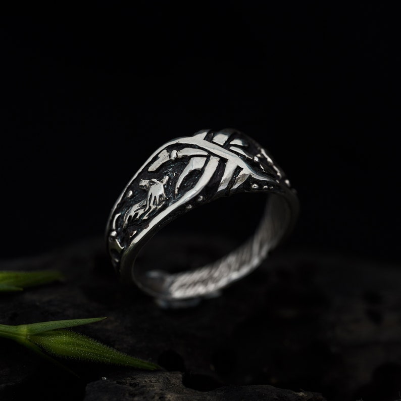 May include: A silver ring with a detailed design featuring a stylized animal figure and intricate lines. The ring has a textured finish and a slightly curved band.