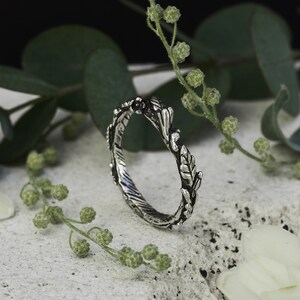 Peut inclure: Une bague argent&eacute;e au design inspir&eacute; de la nature. La bague pr&eacute;sente un anneau textur&eacute; et est orn&eacute;e de d&eacute;tails de feuilles et de branches. La bague est pr&eacute;sent&eacute;e sur une surface blanche avec un feuillage vert en arri&egrave;re-plan.