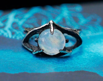 May include: A silver ring with a large, round, white moonstone set in a claw setting. The ring has a unique, eye-like design.