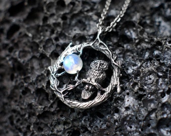 May include: A silver owl pendant necklace with a moonstone and a detailed design of a tree branch. The pendant is hanging on a silver chain.
