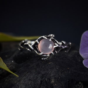 May include: A silver ring with a delicate branch design and a pale pink gemstone. The ring is set on a dark background.