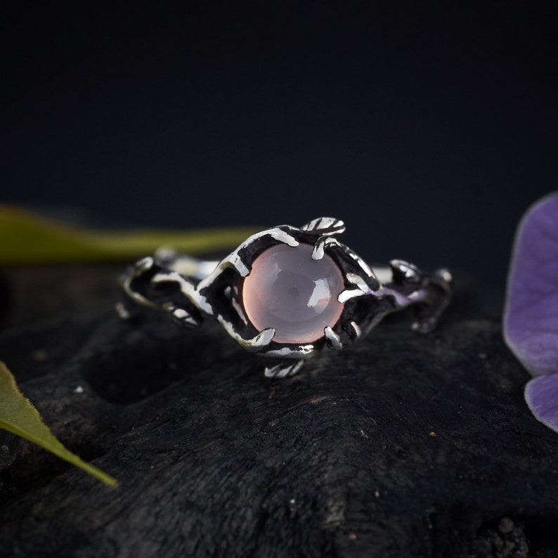 May include: A silver ring with a delicate branch design and a pale pink gemstone. The ring is set on a dark background.