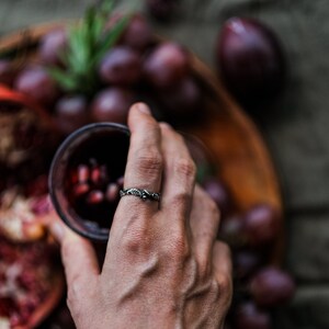 Peut inclure: Une bague en argent avec un motif de feuilles et de baies. La bague est port&eacute;e au doigt, avec un verre de liquide rouge en arri&egrave;re-plan. Des raisins et un bol en bois sont &eacute;galement visibles.