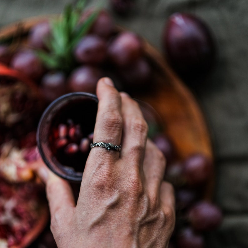 Peut inclure: Une bague en argent avec un motif de feuilles et de baies. La bague est port&eacute;e au doigt, avec un verre de liquide rouge en arri&egrave;re-plan. Des raisins et un bol en bois sont &eacute;galement visibles.