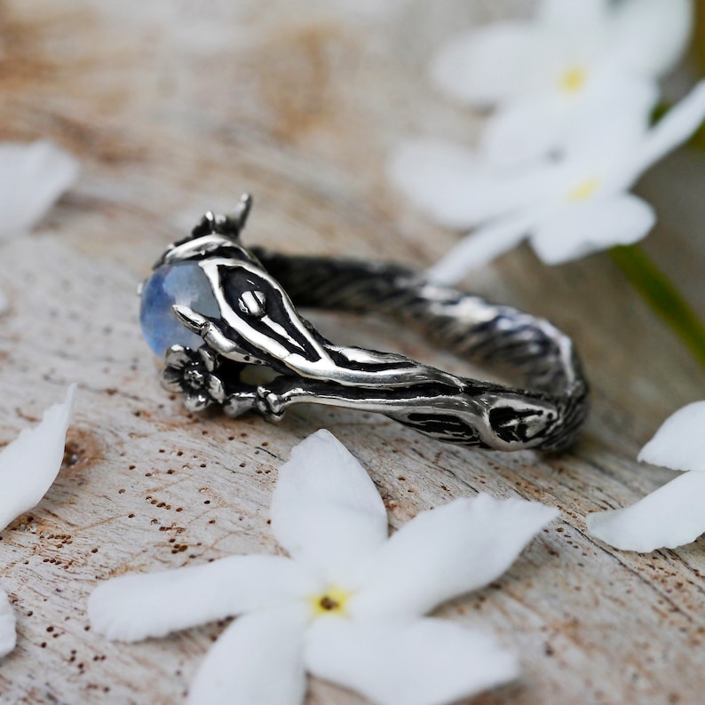 May include: A silver ring with a moonstone set in a branch-like design. The ring is surrounded by white flowers.