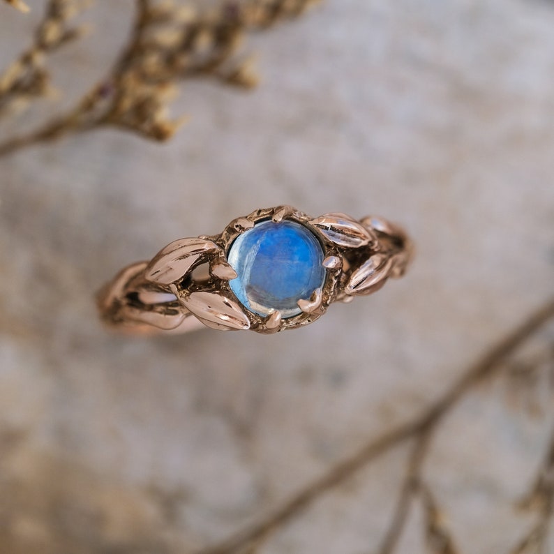 Puede incluir: Un delicado anillo de oro rosa con una piedra de luna azul engastada en un dise&ntilde;o de hojas.