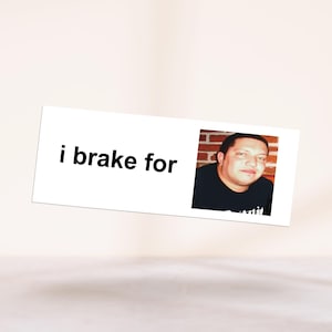 May include: A white rectangular sign with the text "i brake for" and a photo of a man in a black shirt. The man is in front of a brick wall. The sign is slightly angled.