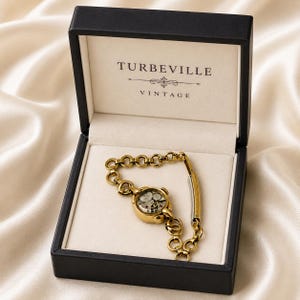May include: A gold-toned watch movement bracelet with a circular exposed gear mechanism and a chain link band, presented inside a black rectangular box with the text TURBEVILLE VINTAGE printed in black on the cream-colored interior lid lining.