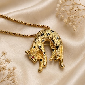 May include: A gold-toned metal pendant shaped like a leaping leopard with a textured surface, black enamel spots, and a small red stone for an eye. The pendant is attached to a gold-toned box chain necklace, creating a cohesive jewelry piece.