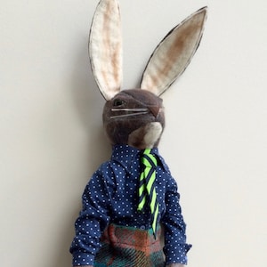 May include: A plush rabbit toy wearing a blue polka dot shirt with a green and black striped tie and plaid trousers. The rabbit has brown fur and long ears.
