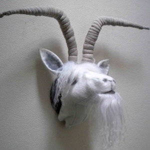 May include: A plush goat head wall mount with long, curved grey horns and a white beard. The goat has a white face with grey accents on the ears and around the eyes. The mount is attached to a wall.