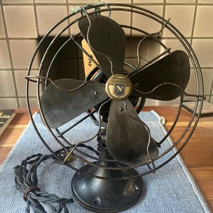 May include: A vintage black metal electric desk fan with a circular wire cage and four dark blades. The center of the fan features a circular emblem with the text "Day-Fan" and "Quality." A black fabric-covered power cord is attached to the base.