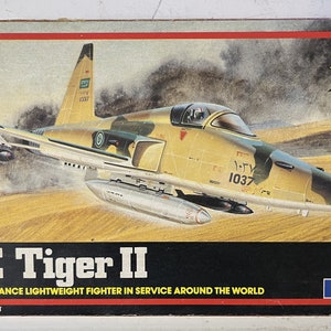 Puede incluir: Un kit de maqueta de un avión de combate F5E Tiger II en un entorno desértico. El avión está pintado con un patrón de camuflaje verde, marrón y beige. La caja del modelo presenta el texto "F5E Tiger II" y "High Performance Lightweight Fighter in Service Around the World".