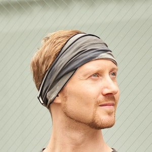 May include: A man wearing a gray t-shirt and a gray and black patterned headband. He is standing in front of a glass wall.