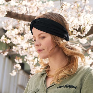 May include: A solid black fabric headband featuring a twisted knot detail at the center front. The headband is worn around the head, resting against the hair, and is constructed from a smooth, flexible material that conforms to the shape of the head.