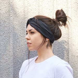 May include: A solid black fabric headband featuring a twisted knot detail at the front, worn across the forehead. The headband is made of a smooth, flexible material that sits securely around the head, providing a clean and simple accessory.