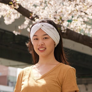 May include: A white twisted headband made of fabric worn on the head, paired with a plain mustard yellow short-sleeved v-neck t-shirt. The headband features a central knot detail and a soft, ribbed texture, providing a simple accessory.