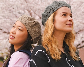 May include: Two women wearing crocheted berets, one in brown and one in gray, stand back to back with their heads tilted up. They are both wearing casual clothing and are standing in front of a blurred background of pink cherry blossoms.