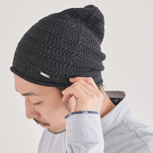 May include: A dark charcoal gray knit beanie featuring a textured, repeating wave-like pattern throughout the fabric. The beanie includes a small, rectangular silver-toned metal tag attached to the lower edge, engraved with the text "Charm".