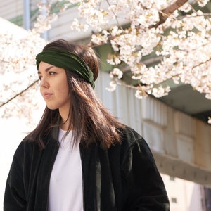 May include: A dark olive green fabric headband featuring a twisted knot detail at the front. The headband is made of a soft, flexible material that sits securely across the forehead, providing a simple and functional hair accessory design.