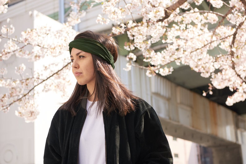 May include: A dark olive green fabric headband featuring a twisted knot detail at the front. The headband is made of a soft, flexible material that sits securely across the forehead, providing a simple and functional hair accessory design.