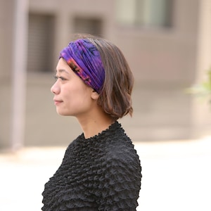Puede incluir: Una mujer con una diadema tie-dye morada y naranja. La diadema est&aacute; enrollada alrededor de la cabeza. Lleva una blusa negra texturizada. El fondo est&aacute; borroso. La foto est&aacute; tomada al aire libre.