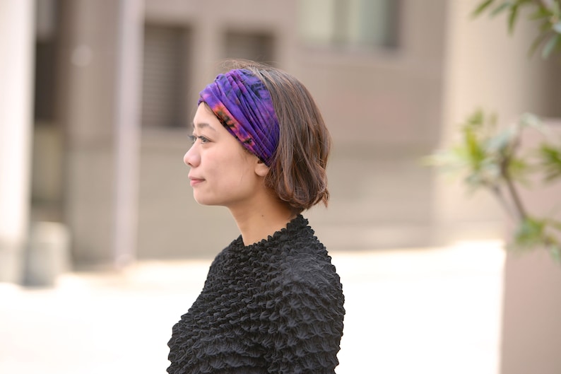 Puede incluir: Una mujer con una diadema tie-dye morada y naranja. La diadema est&aacute; enrollada alrededor de la cabeza. Lleva una blusa negra texturizada. El fondo est&aacute; borroso. La foto est&aacute; tomada al aire libre.