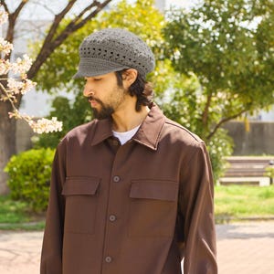 May include: A gray crocheted newsboy cap with a brim. The cap is worn with a brown button-up shirt with two chest pockets. The shirt is worn over a white t-shirt. The background includes trees and a park setting.