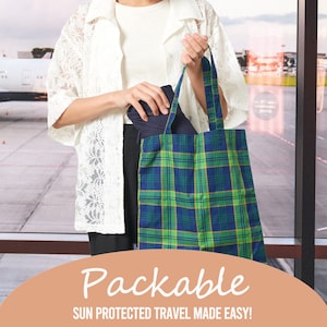 Puede incluir: Una mujer sostiene una bolsa de mano de lona a cuadros verde y azul. La bolsa es plegable y tiene el texto "Packable Sun Protected Travel Made Easy!"
