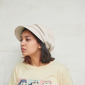May include: A beige bucket hat with a rounded crown and a short, downward-sloping brim. The hat is worn by a person wearing a light yellow t-shirt with a colorful graphic print. The background is a white wall.