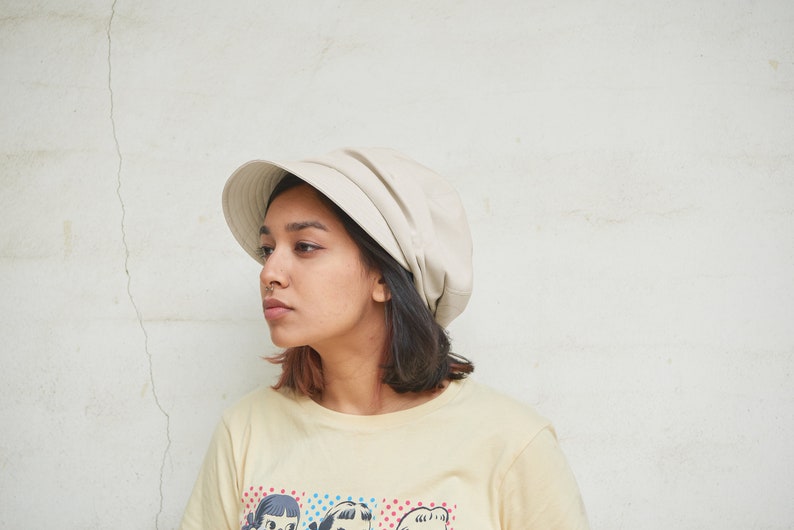 May include: A beige bucket hat with a rounded crown and a short, downward-sloping brim. The hat is worn by a person wearing a light yellow t-shirt with a colorful graphic print. The background is a white wall.