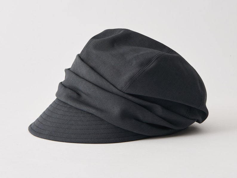 May include: A black newsboy hat with a structured brim and a gathered fabric crown. The hat has a classic design and is made of a solid, dark fabric. The brim is slightly curved and has visible stitching.