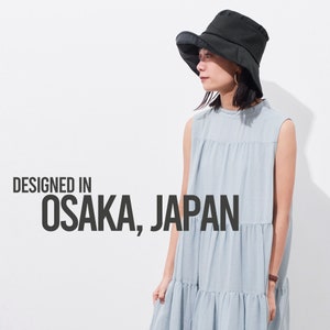 May include: A woman wearing a light blue sleeveless dress and a black bucket hat. The text "DESIGNED IN OSAKA, JAPAN" is printed on a white background.