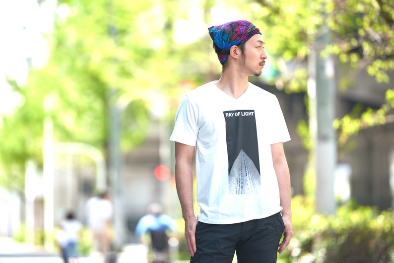 Puede incluir: Una camiseta blanca con cuello en V con el texto "RAY OF LIGHT" sobre un gr&aacute;fico arquitect&oacute;nico en blanco y negro. La persona lleva un gorro tie-dye colorido y pantalones negros.