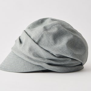 May include: A light grey newsboy cap with a curved peak. The hat features a soft, draped design and is made from a textured fabric. This cap is a stylish accessory for everyday wear.