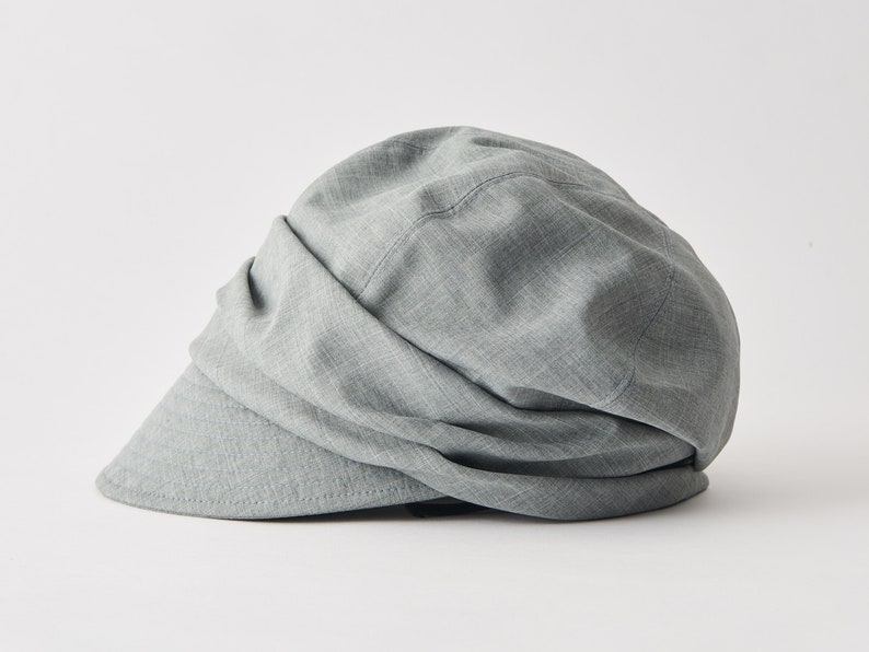 May include: A light grey newsboy cap with a curved peak. The hat features a soft, draped design and is made from a textured fabric. This cap is a stylish accessory for everyday wear.