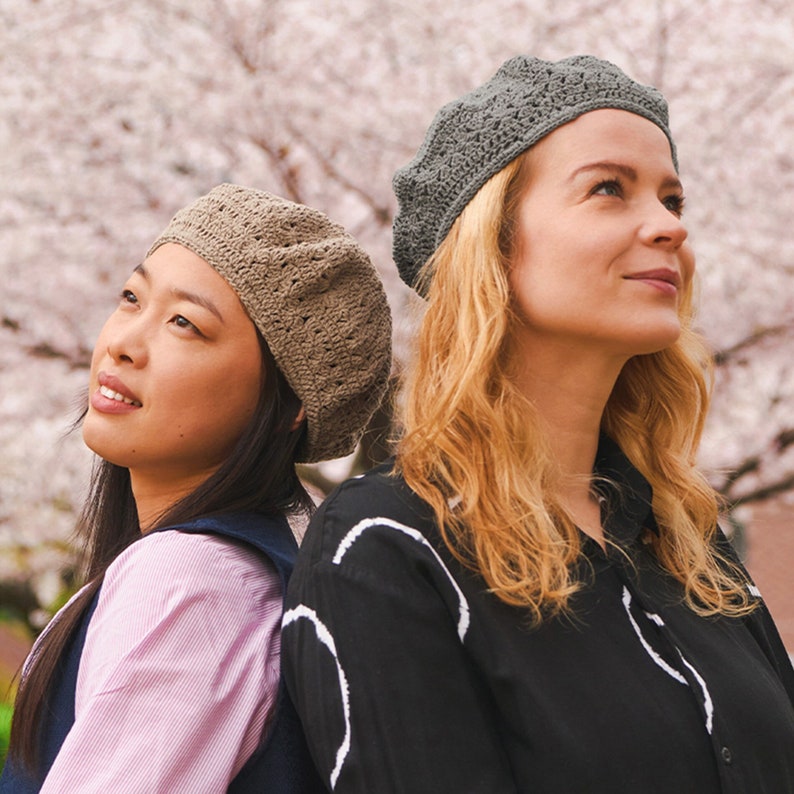 以下が含まれることがあります： 2人の女性が、茶色と灰色のクロシェのベレー帽をかぶって、背中合わせに立っています。2人ともカジュアルな服装で、ピンクの桜の花がぼやけて見える背景の前に立っています。