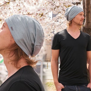 Pode incluir: Uma pessoa usando um gorro cinza e uma camiseta preta de gola em V. A pessoa est&aacute; em p&eacute; na frente de uma &aacute;rvore com flores brancas.
