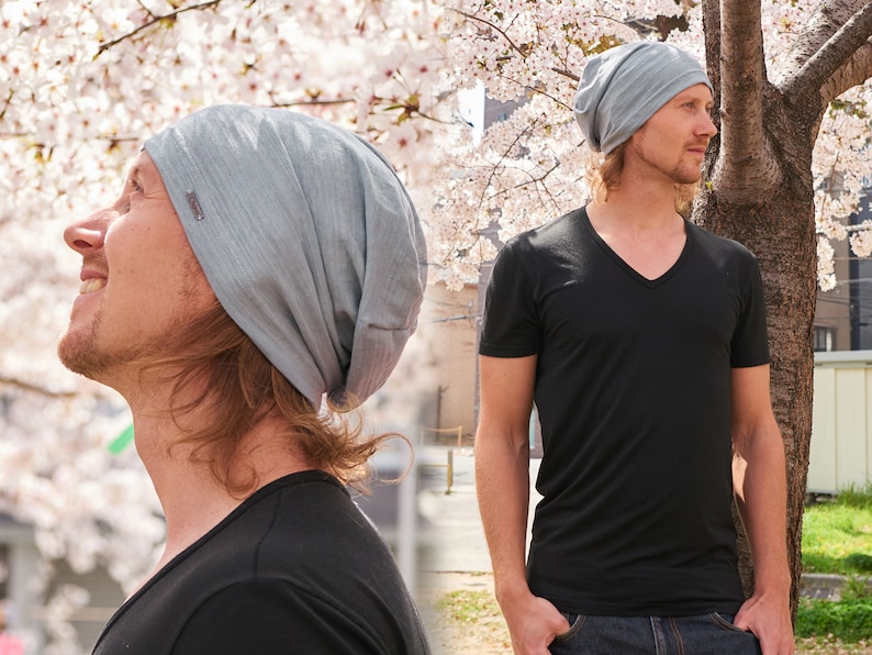 Pode incluir: Uma pessoa usando um gorro cinza e uma camiseta preta de gola em V. A pessoa est&aacute; em p&eacute; na frente de uma &aacute;rvore com flores brancas.