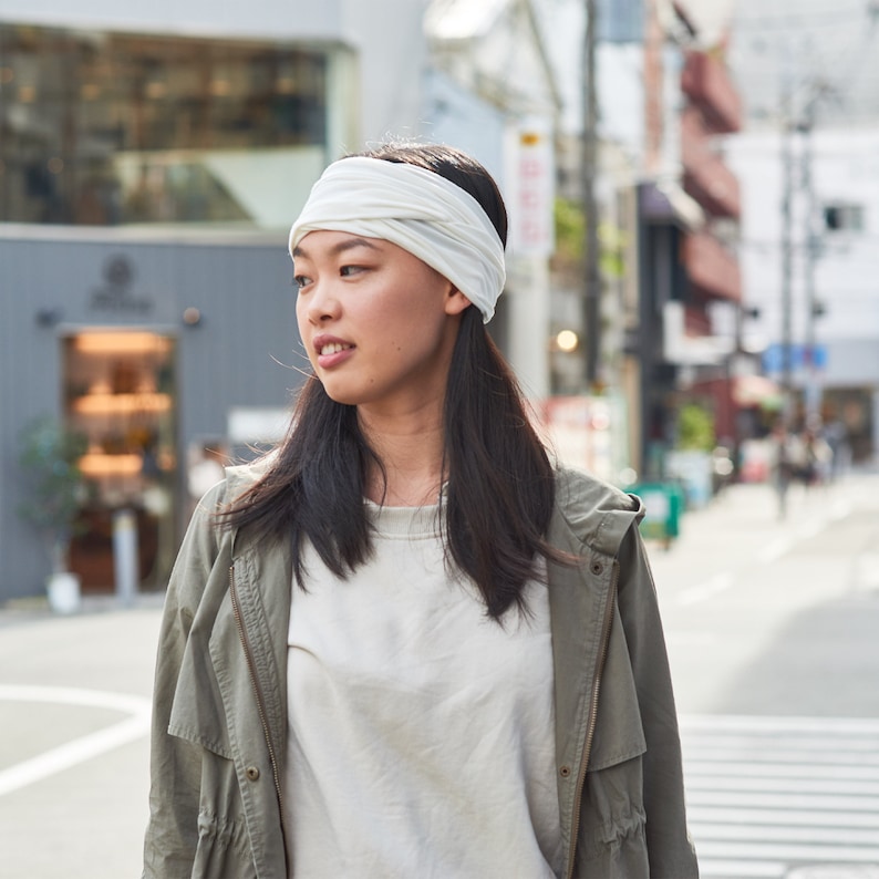 May include: A woman with long black hair is wearing a white headband and a green jacket. She is standing in a city street.