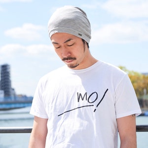 Pode incluir: Um homem usando um gorro cinza e uma camiseta branca com a palavra "MO!" escrita em preto. Ele est&aacute; em p&eacute; em frente a um corpo de &aacute;gua com uma ponte ao fundo.