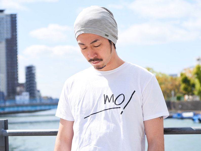Pode incluir: Um homem usando um gorro cinza e uma camiseta branca com a palavra "MO!" escrita em preto. Ele est&aacute; em p&eacute; em frente a um corpo de &aacute;gua com uma ponte ao fundo.