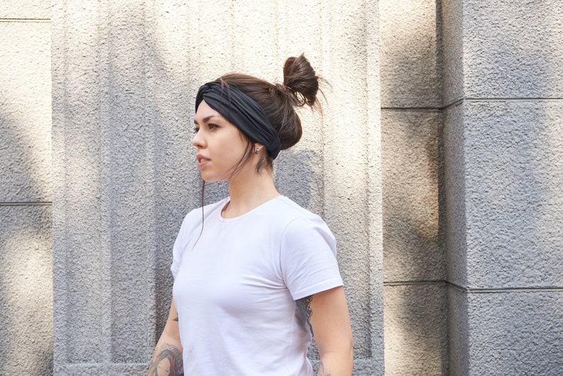 May include: A solid black fabric headband featuring a twisted knot detail at the front, worn across the forehead. The headband is made of a smooth, flexible material that sits securely around the head, providing a clean and simple accessory.