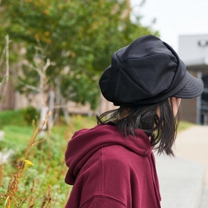 K&ouml;nnte beinhalten: Eine Person tr&auml;gt eine schwarze Newsboy-M&uuml;tze mit einem weinroten Kapuzenpullover.