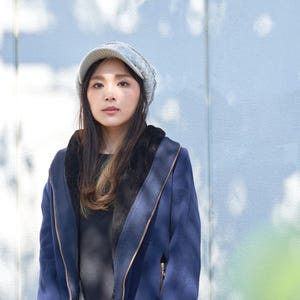 May include: A woman wearing a blue jacket with a gray knitted hat. The jacket has a zipper and a hood. The woman is standing in front of a light blue wall.